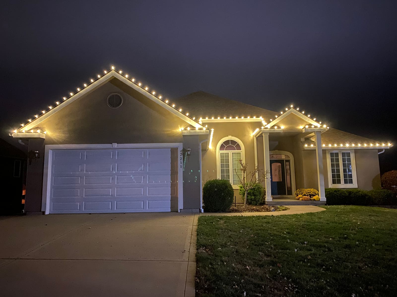 Stucco ranch home with warm white Christmas lights on the garage peak, roofline and covered porch